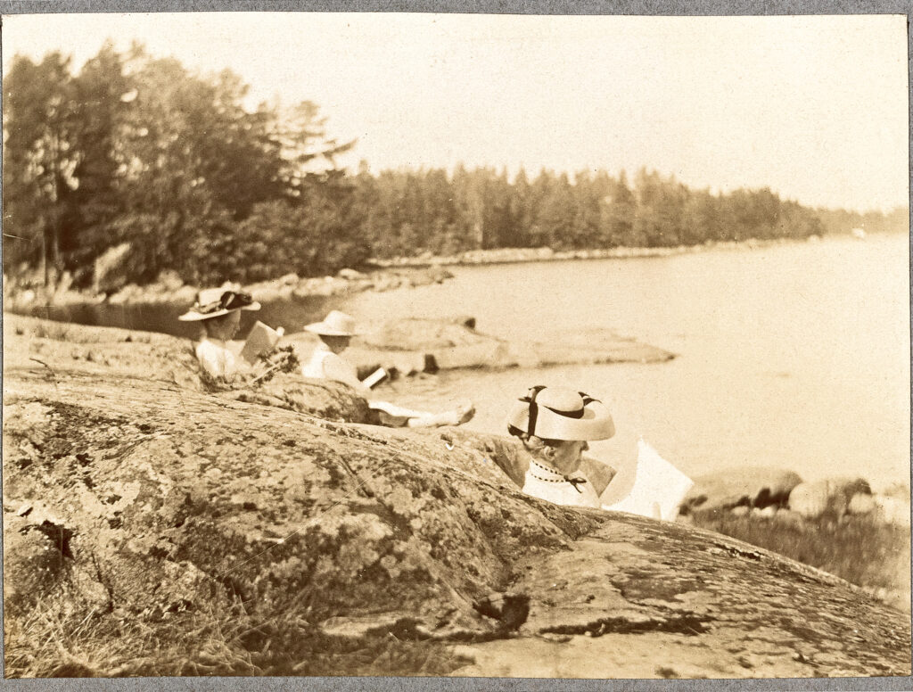 Svartvitt foto av tre kvinnor i hattar som sitter på klippor vid en sjö, omgivna av träd och en klippig strandlinje i bakgrunden.