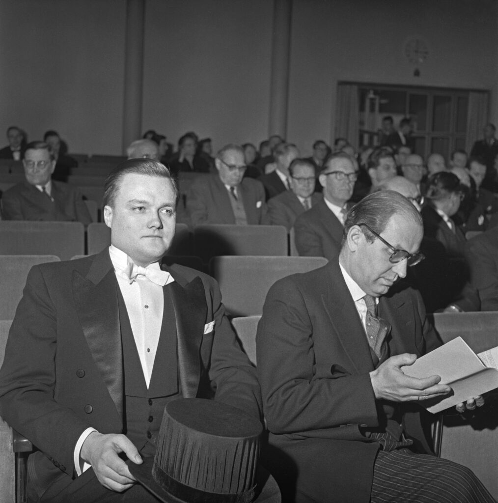 Svartvitt foto av två män i kostym i en auditorium. Den yngre mannen till vänster håller en studentmössa, medan den äldre mannen till höger läser ett programblad. Flera personer syns i bakgrunden.