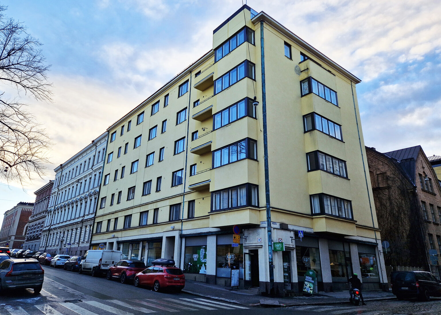 Färgfoto av ett gult funkishus med stora fönster i hörnläge, butikslokaler i bottenplan, bilar parkerade längs gatan och blå himmel med moln.