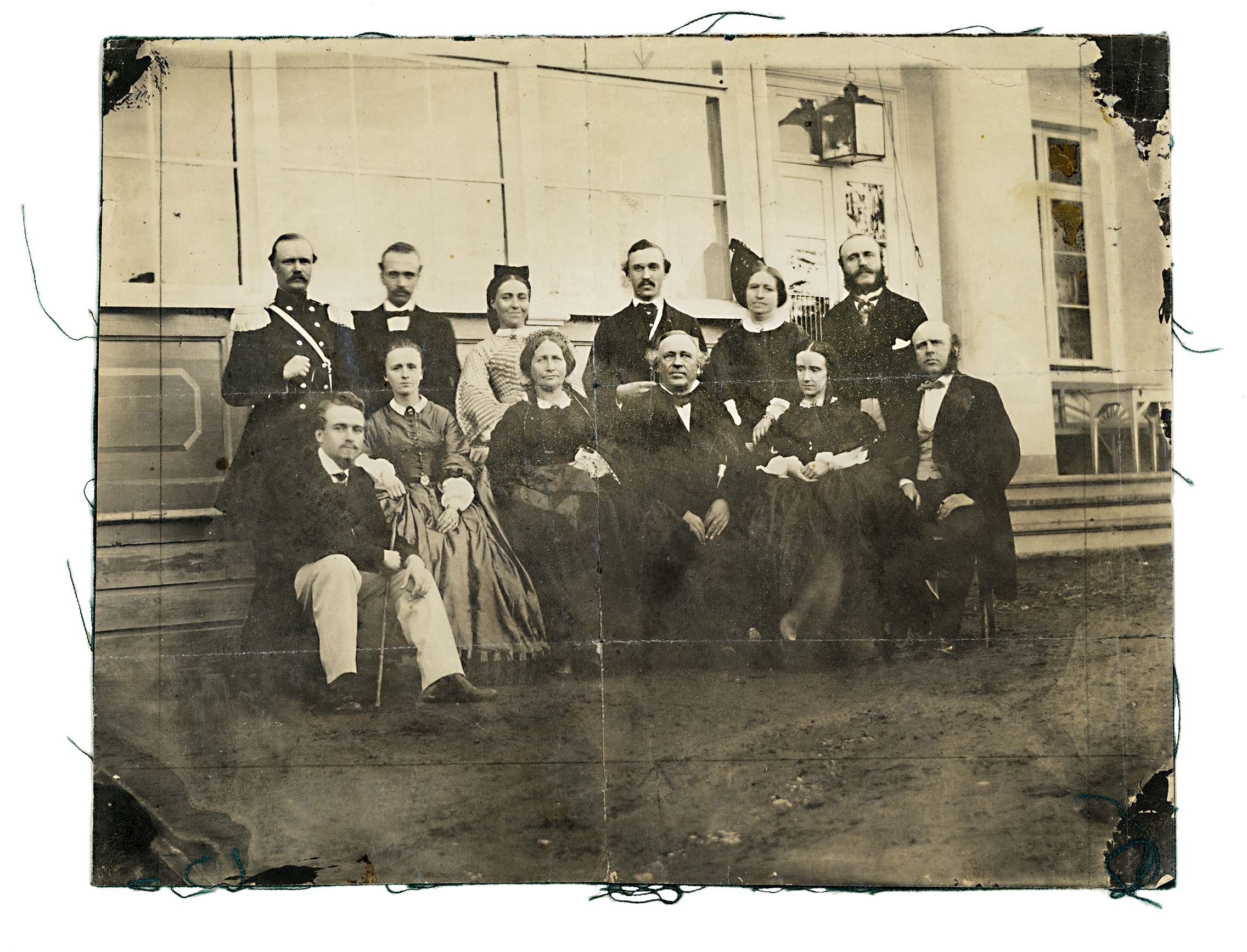 Svartvitt foto av tolv personer, män och kvinnor i formella 1860-talskläder, sittande och stående i två rader framför ett hus med stora fönster och veranda.