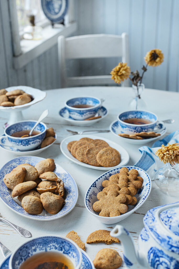 Foto av ett ljust dukat bord med blåvitt porslin, koppar med te, fat med pepparkakor och småbröd samt torkade blommor i små vaser i lantlig miljö.