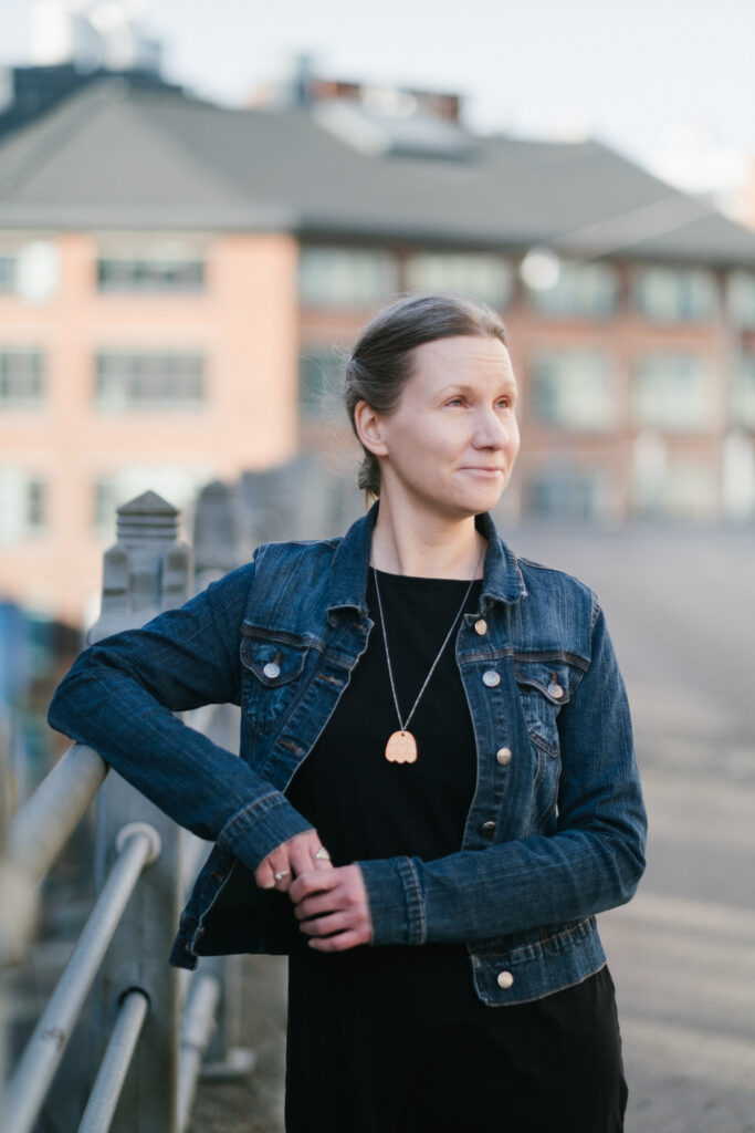 Färgfoto av kvinna i jeansjacka och svart klänning, stående vid ett räcke utomhus, med suddig byggnad i bakgrunden, blickande snett uppåt.