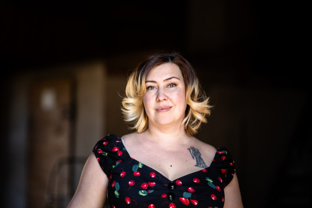 Färgfoto av kvinna med axellångt blont hår, klänning med körsbärsmönster och tatuering på bröstet, stående mot mörk och suddig bakgrund.